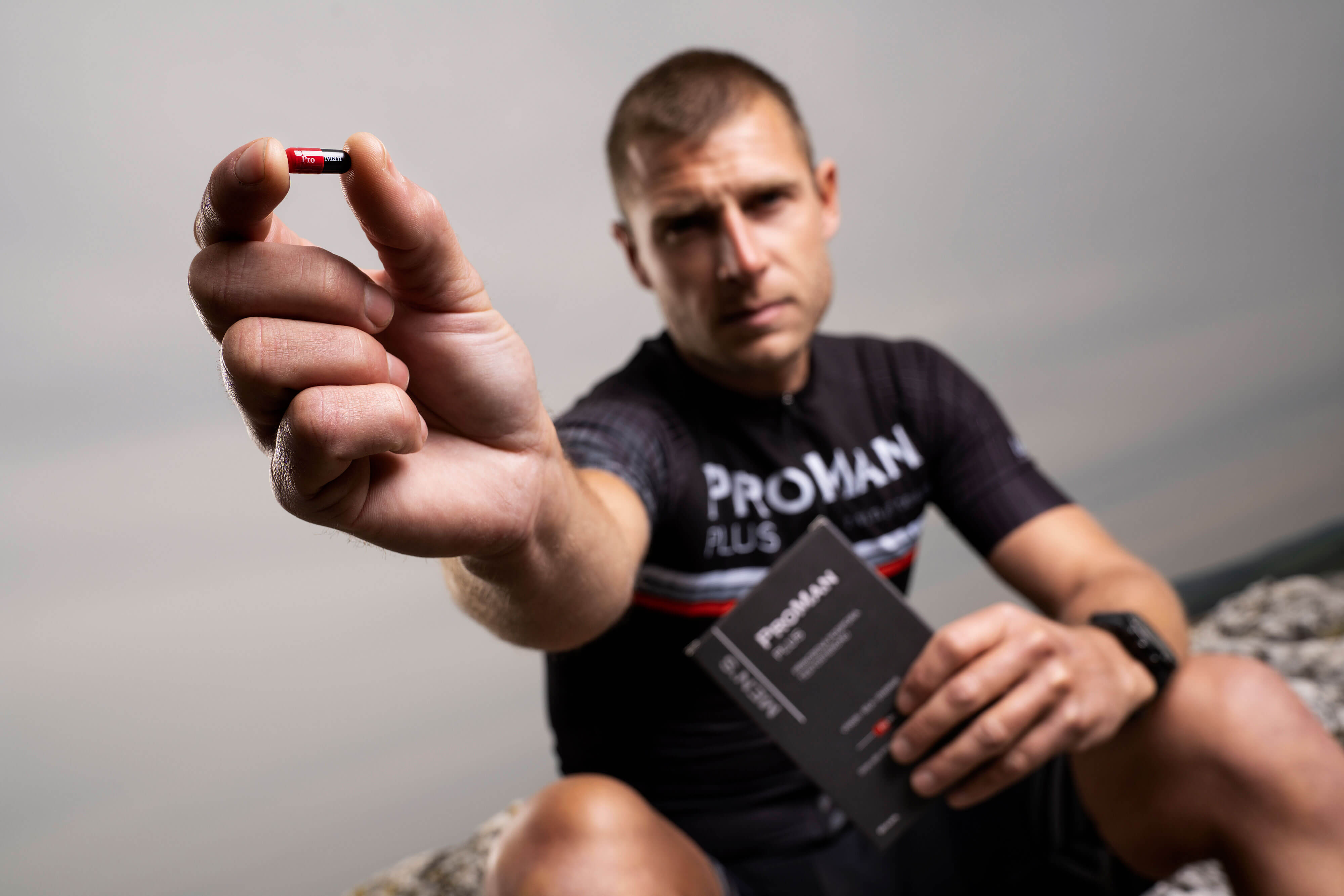 Natural Pharmaceuticals ProMan® Plus Egy férfi „ProMan® Plus” sportpólóban egy étrend-kiegészítő kapszulát tart a lencse felé nyújtva. Másik kezében a termék csomagolását tartja. Elmosódott, szabadtéri háttér.